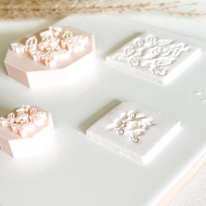 May include: Four craft stamps with floral designs. Two stamps are hexagonal and pink, while the other two are square and white. The stamps have raised floral patterns, ideal for adding decorative elements to craft projects.