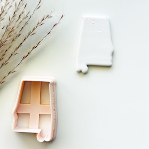 May include: A light pink plastic cookie cutter shaped like the state of Alabama, positioned next to a white clay cutout of the same shape. The cookie cutter features a cross-shaped internal support structure and a smooth, molded plastic edge.