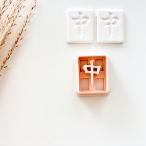 May include: Three Mahjong tile cookie cutters. Two white tiles with a Chinese symbol are above a peach-colored cutter with the same symbol. The cutters are on a white surface with a dried plant stem in the upper left corner.