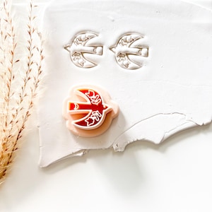 May include: A red and white bird-shaped clay cutter and two bird-shaped impressions. The cutter is red with white details and a light pink outline. The impressions are detailed with leaf-like patterns.