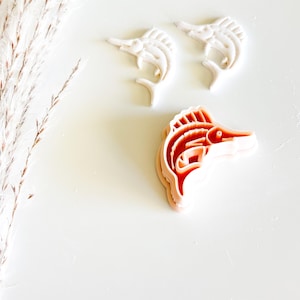 May include: Three plastic cookie cutters in the shape of a marlin fish. Two are white, and one is peach-colored. The cutters are on a white surface with a dried plant stem in the upper left corner.