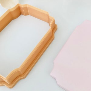 May include: A beige plastic cookie cutter in the shape of a rectangle with rounded corners and a decorative border. A light pink rectangle of fondant is next to the cutter.