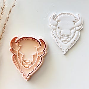 May include: Two bison-shaped cookie cutters. One is a light peach color with a darker outline, and the other is white. Both cutters have detailed features of the bison's face and horns. The cutters are likely used for baking or crafting.