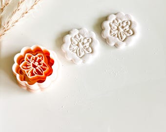 May include: A flower-shaped cookie cutter and two flower-shaped cookies. The cookie cutter is orange and has a detailed floral design. The cookies are white and have a similar floral pattern. The items are on a white surface.