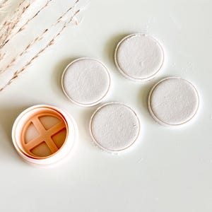 May include: A light peach-colored cookie cutter with a cross-shaped interior, next to four round, light-colored cookies. The cookies have a slightly textured surface and a defined edge. The items are arranged on a white surface.