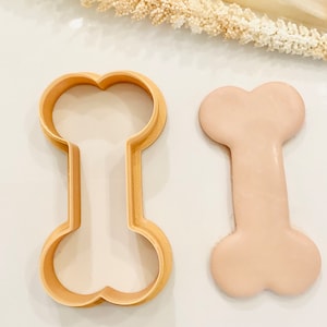 Peut inclure: Un emporte-pièce en plastique marron en forme d'os de chien à côté d'un biscuit en forme d'os de couleur marron clair.