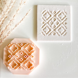 May include: A light pink, octagonal cookie stamp with a white geometric pattern next to a white square with the same pattern. The stamp is likely used for creating decorative designs on cookies or other baked goods.