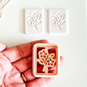 May include: A floral cookie cutter with a rectangular frame, featuring a flower design in orange and white. Two white, rectangular cookies with embossed floral patterns are also visible. The items are on a white surface.