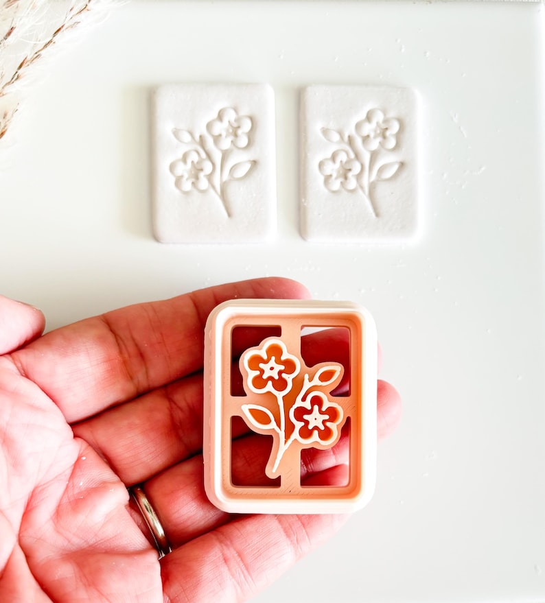 May include: A floral cookie cutter with a rectangular frame, featuring a flower design in orange and white. Two white, rectangular cookies with embossed floral patterns are also visible. The items are on a white surface.
