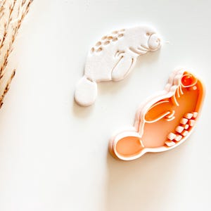 May include: Two manatee-shaped cookie cutters. One is white, showing the manatee's form with textured details. The other is orange, with a detailed interior, and a light pink outline. The cutters are on a white surface.