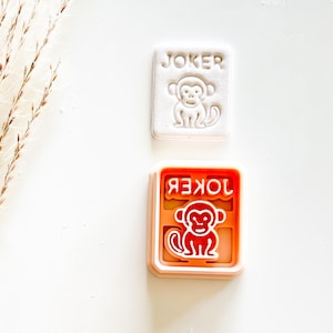 May include: Two cookie cutters. One is white with the word "JOKER" and a monkey design. The other is orange with the word "JOKER" in reverse and a monkey design. Both are rectangular.