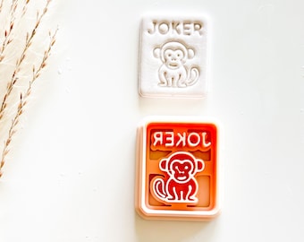 May include: Two cookie cutters. One is white with the word "JOKER" and a monkey design. The other is orange with the word "JOKER" in reverse and a monkey design. Both are rectangular.