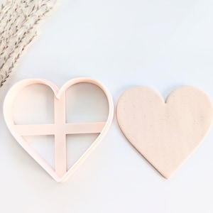 May include: A pink heart-shaped cookie cutter with a cross-shaped design inside. The cutter is next to a plain pink heart-shaped cookie.