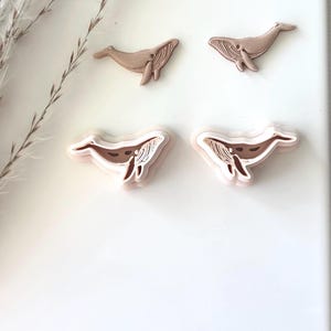 May include: Four whale-shaped cookie cutters, two large and two small. The cutters are made of a light brown plastic and are arranged on a white background.