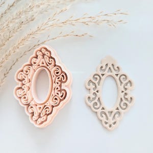 May include: Two pink plastic cookie cutters in the shape of an ornate oval frame. The cutter on the left is a solid shape, while the cutter on the right is an outline.