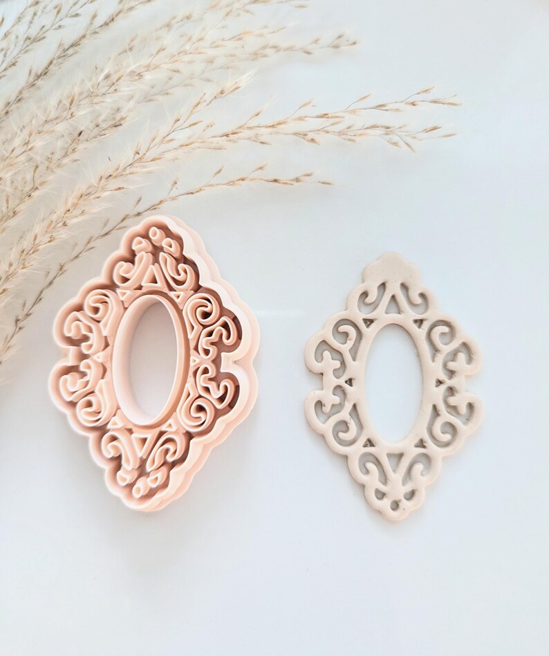 May include: Two pink plastic cookie cutters in the shape of an ornate oval frame. The cutter on the left is a solid shape, while the cutter on the right is an outline.