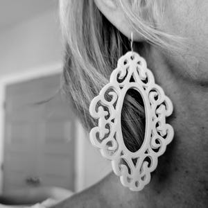 May include: White filigree earrings with a large oval cutout in the center. The earrings are made of a lightweight material and have a delicate, feminine design.
