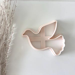 May include: A light pink dove-shaped cookie cutter. The cutter is made of plastic and has a cross-shaped support inside the dove's body. The dove is in flight, with its wings spread. The cutter is on a white surface.