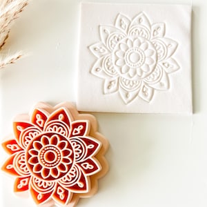 May include: A floral-shaped cookie cutter with a detailed design, next to a square impression of the same design. The cutter is a layered design in shades of orange and white. The square impression is white.