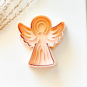 May include: A light peach-colored angel-shaped cookie cutter. The cutter has a layered design with detailed wings, a halo, and a flowing gown. The cutter is made of plastic and is designed for baking. The angel is centered on a white background.