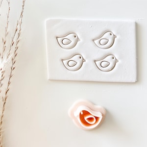 May include: A white rectangular mold featuring four bird-shaped cutouts, alongside a separate bird-shaped cutter. The bird designs have a simple outline with a circular eye detail. The cutter is pink and orange.
