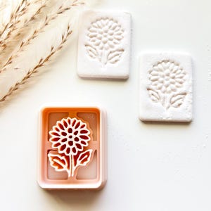 May include: A pink rectangular cookie cutter with a flower design, next to two white rectangular cookies with the same flower imprint. The cookies and cutter are on a white surface.