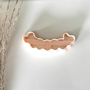May include: A light peach-colored cookie cutter in the shape of a cloud with a horizontal bar across the center. The cutter has a wavy bottom edge and a smooth top edge. The cutter is made of plastic.