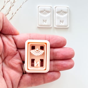 May include: A light peach-colored rectangular cookie cutter with a decorative design and the letter "N" is held in a hand. Two white rectangular stamps with the same design and letter are also visible.