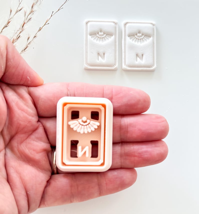 May include: A light peach-colored rectangular cookie cutter with a decorative design and the letter "N" is held in a hand. Two white rectangular stamps with the same design and letter are also visible.