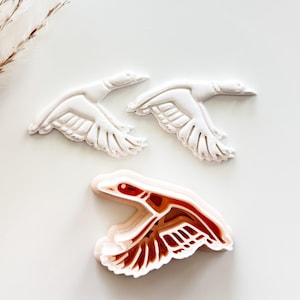 May include: Three white and brown duck-shaped cookie cutters. Two are solid white, and one is white with brown and red details. The ducks are in flight, with wings spread. The cutters are on a white surface.