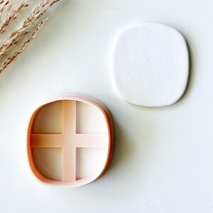 May include: A light peach-colored square cookie cutter with rounded corners and a cross-shaped internal divider, positioned next to a smooth, flat, white square piece of clay with rounded corners that matches the shape of the cutter.