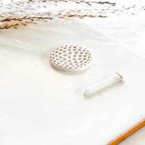 Pu&ograve; includere: Un set bianco in ceramica composto da un segnaposto per pallina da golf e un tee. Il segnaposto &egrave; rotondo con un motivo a nido d'ape. Il tee ha un design allungato. Il set &egrave; esposto su una superficie bianca.