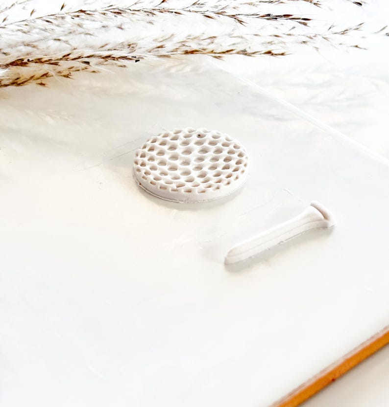Pu&ograve; includere: Un set bianco in ceramica composto da un segnaposto per pallina da golf e un tee. Il segnaposto &egrave; rotondo con un motivo a nido d'ape. Il tee ha un design allungato. Il set &egrave; esposto su una superficie bianca.
