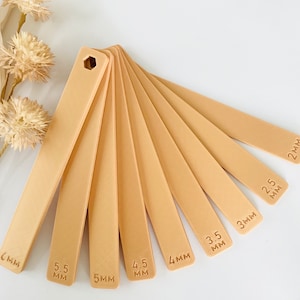 May include: A set of ten beige plastic rulers with measurements in millimeters, ranging from 2 mm to 6 mm. The rulers are arranged in a fan shape, with the smallest ruler at the top and the largest ruler at the bottom.