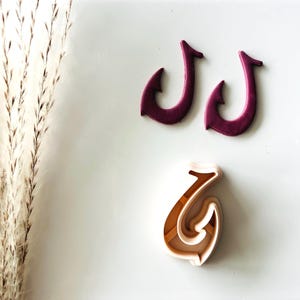 May include: Three hook-shaped items. Two are a deep purple color, and the third is a light tan color. The items are arranged on a white surface. The hooks have a curved design with a pointed end.