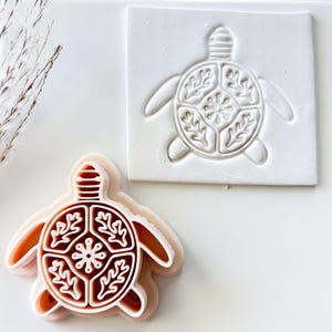 May include: A brown and white turtle-shaped cookie cutter with a detailed design, next to a white square with the same turtle design. The turtle design includes leaf and floral patterns.