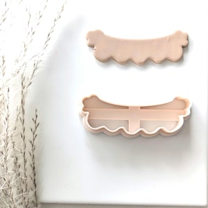May include: Two plastic cookie cutters in the shape of a scalloped edge banner. The top cutter is solid and the bottom cutter is open.