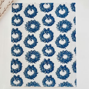 May include: White paper with a repeating pattern of blue wreaths. Each wreath is detailed with leaves and a bow. The design is evenly spaced across the paper, creating a classic and festive look. The paper is likely used for wrapping or crafting.