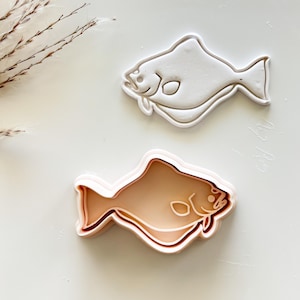 May include: Two fish-shaped cookie cutters. One is white, the other is light brown. Both have a detailed outline of a fish with fins, eyes, and gills. These cutters are ideal for creating fish-themed cookies or treats.