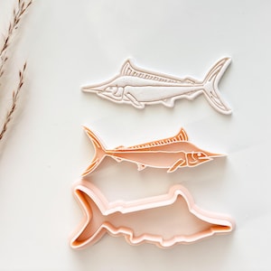 May include: Three marlin-shaped cookie cutters in varying shades of pink and peach. The top cutter is a light pink, the middle one is peach, and the bottom one is a darker pink outline. The cutters are arranged on a white surface.