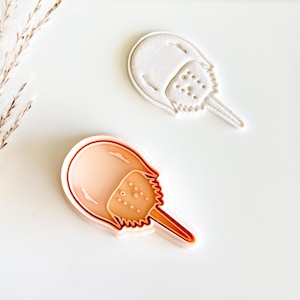 May include: Two horseshoe crab cookie cutters. One is a light peach color with a darker outline, and the other is white. The cutters are on a white surface. The horseshoe crab design is detailed with a segmented body and tail.