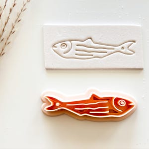 May include: A fish-shaped cookie cutter and a matching impression stamp. The cutter is orange and white, while the stamp is a light beige. The fish design features a detailed eye, gills, and tail.