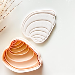 May include: A seashell-shaped cookie cutter in two parts. One part is a light peach color, and the other is white. The cutter is designed to create seashell-shaped cookies or pastries. The design features the curved lines of a seashell.