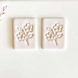 May include: Two rectangular, off-white ceramic tiles with a floral design. Each tile features an embossed image of a flower with three blossoms and leaves. The design is centered on the tile, creating a symmetrical appearance. The tiles are set against a white background.