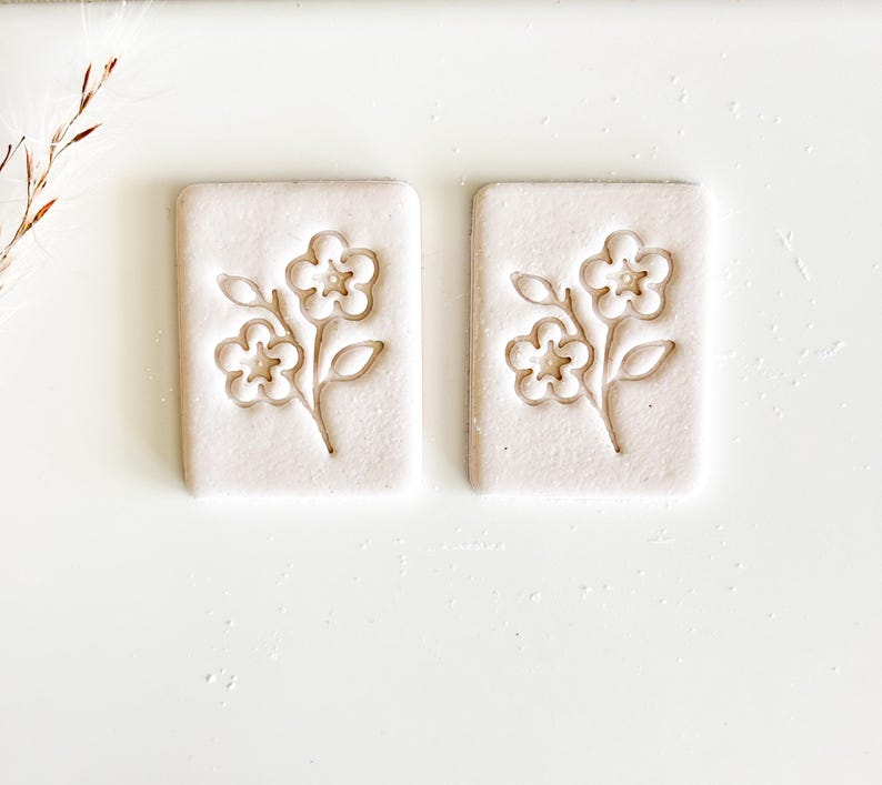 May include: Two rectangular, off-white ceramic tiles with a floral design. Each tile features an embossed image of a flower with three blossoms and leaves. The design is centered on the tile, creating a symmetrical appearance. The tiles are set against a white background.