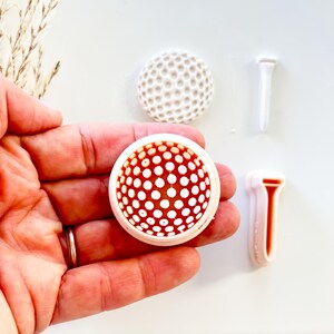 May include: A hand holding a golf ball-shaped object with a white border and a red interior dotted with white circles. Two white golf tees and a white golf ball are also visible.