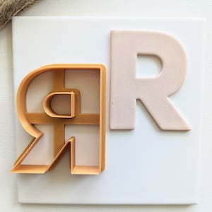 May include: A close-up of a cookie cutter and a piece of clay, both shaped like the letter "R". The cookie cutter is gold and the clay is a light pink color.