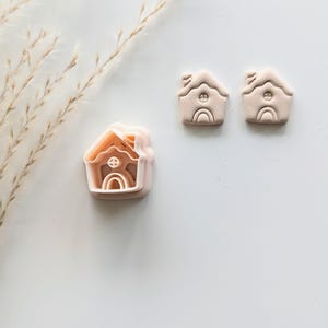 May include: Three pink plastic cookie cutters shaped like houses with a rainbow and window detail. The cutters are on a white surface.