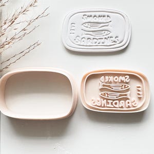 May include: A set of three oval-shaped cookie cutters and stamps in light peach and white. The design features two fish and the text SMOKED SARDINES. One piece is a plain oval cutter, one is a stamp with the fish design, and one is a combined cutter.