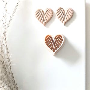 May include: Three heart-shaped cookie cutters, two are solid and one is open, all with a ribbed design. The cutters are a light brown color and are arranged on a white surface.